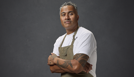 Tony Sokraa against a dark grey background, wearing an apron, cross armed staring into the camera
