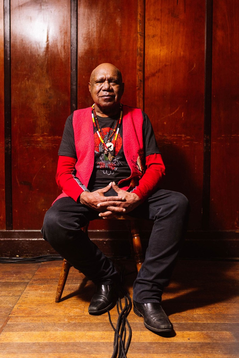 musican-archie-roach-against-a-wooden-panel-wall-in-a-red-vest