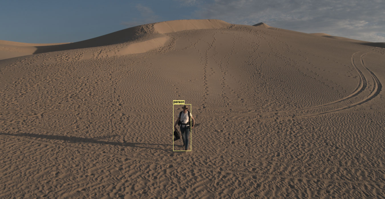 man-leaves-footprints-as-he-walks-across-a-sand-dune-with-camera-gear-a-yellow-box-surrounds-his-body-indicating-artificial-intelligence-is-watching-him-and-identifying-his-shape-as-a-person