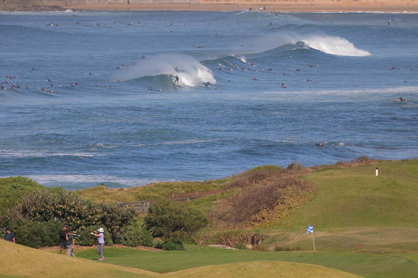  golfers-on-rolling-hills-in-foreground-many-surfers-on-olling-waves-in-background