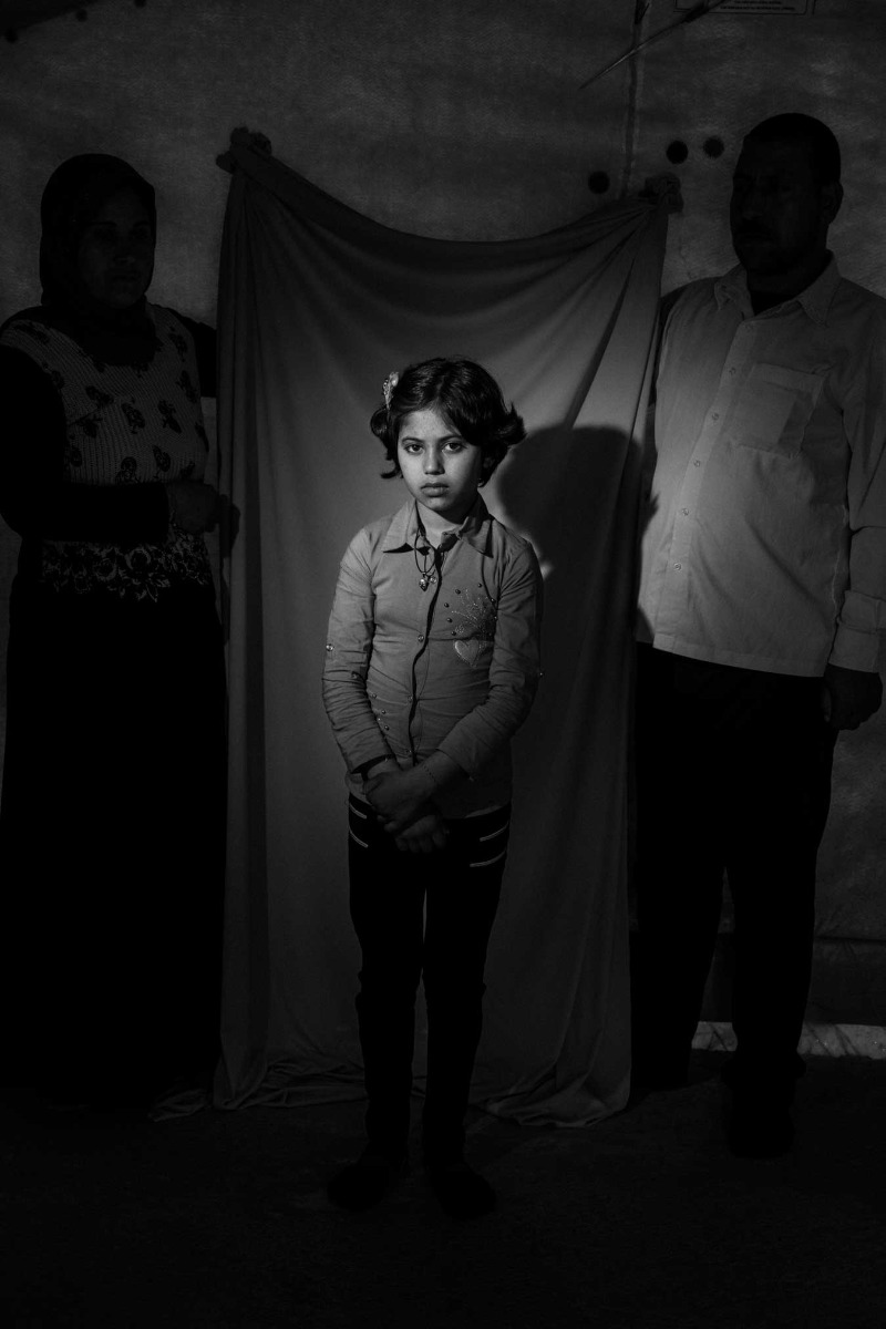 black-and-white-image-of-young-girl-with-pigtails-two-adults-stand-behind-her-holding-up-a-background