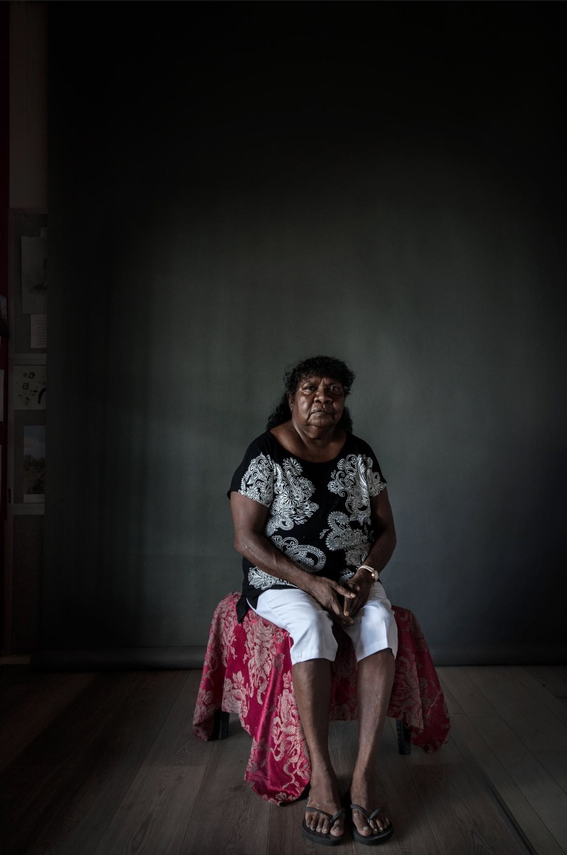 indigenous-australian-woman-casually-dressed-sits-on-a-stool-covered by-red-fabric-against-a-grey-background