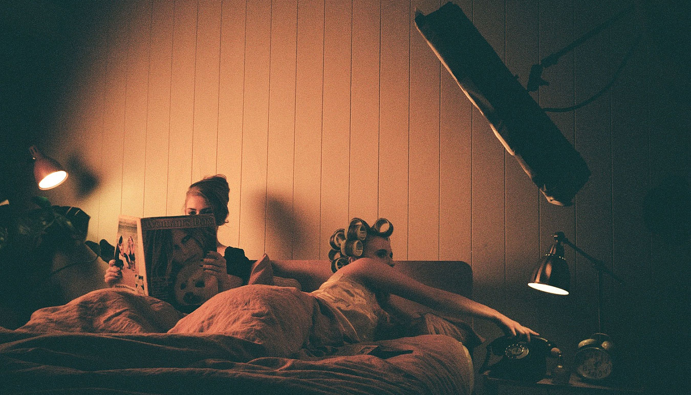 a-behind-the-scenes-image-shot-on-grainy-film-two-women-in-bed-one-reading-a-book-the other-hair-in-rollers-is-reaching-to-answer-a-phone-with-film-lighting-above